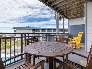 A wooden deck with a round table and chairs, overlooking a swimming pool and a building with a blue roof. - Bay View Villas 203 | Waterfront, Community Pool (Tybee Island)