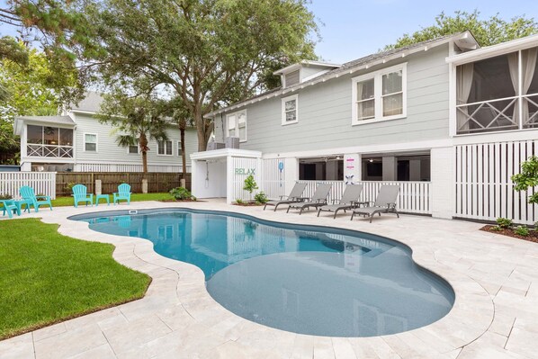 A swimming pool with a curved edge, surrounded by a patio with chairs and a small lawn. - Butler Beach Cottage | Pet-Friendly, Quiet Neighborhood (Tybee Island)