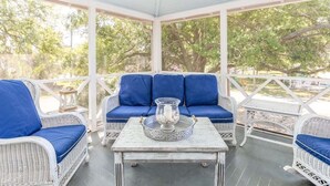 A covered porch with wicker furniture and blue cushions, a metal tray with a candle, and a view of trees. - Butler Beach Cottage | Pet-Friendly, Quiet Neighborhood (Tybee Island)