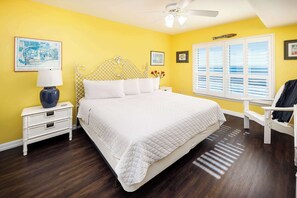 A bedroom with a white bed, a nightstand with a lamp, a chair, and a window with blinds.
