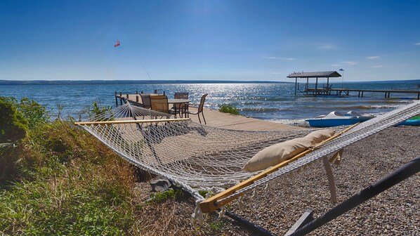 A hammock on a pebbled beach with a view of the water and a dock. - A Shore Thing: Breathtaking Panoramic Views and Spectacular Design Await You! (Varick)