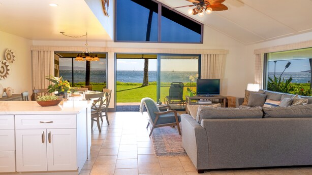 Una sala de estar moderna con un sofá grande de color gris, un comedor con mesa y sillas, una cocina con armarios blancos y ventanas amplias que ofrecen vistas al mar.