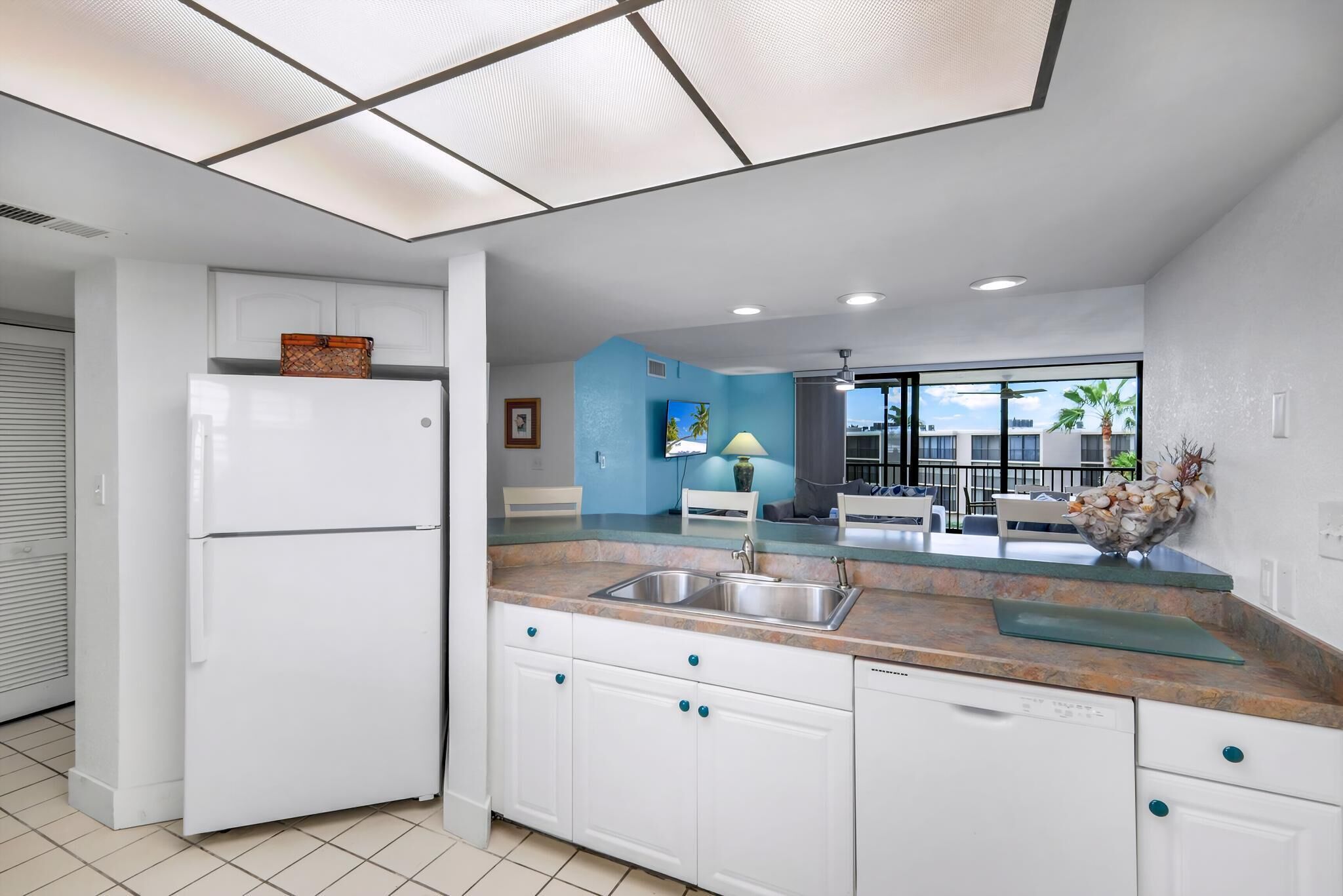 A modern kitchen with white cabinets, a double-door refrigerator, a granite countertop, and a large window offering a view of the outdoors.