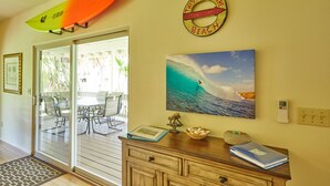 A room with a glass door leading to an outdoor area, a surfboard mounted on the wall, a beach-themed sign, a painting of a surfer, and a wooden cabinet with decorative items. - Hawaiian plantation-style home, perfect for families.  (Hauula)