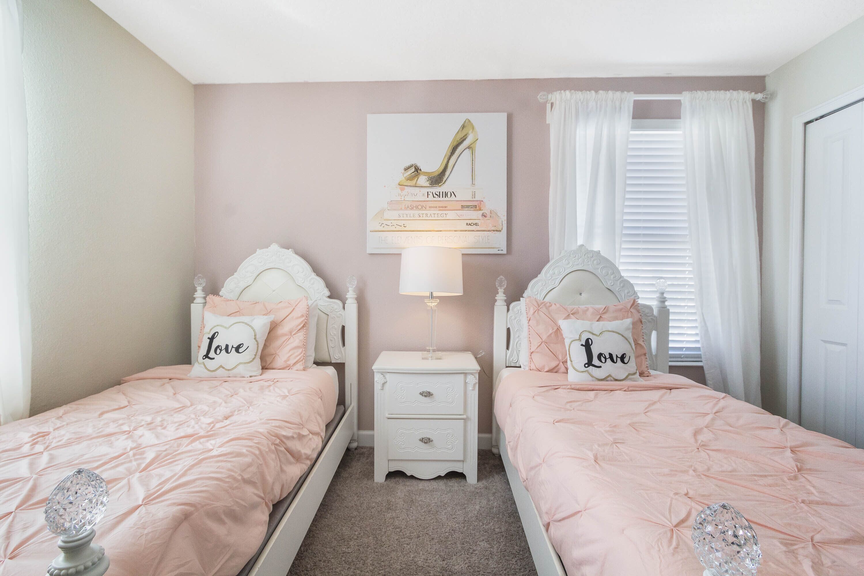 Une chambre avec deux lits, chacun recouvert d’une couvre-lit rose et agrémenté d’oreillers assortis. Une table de chevet avec une lampe se trouve entre les deux lits.