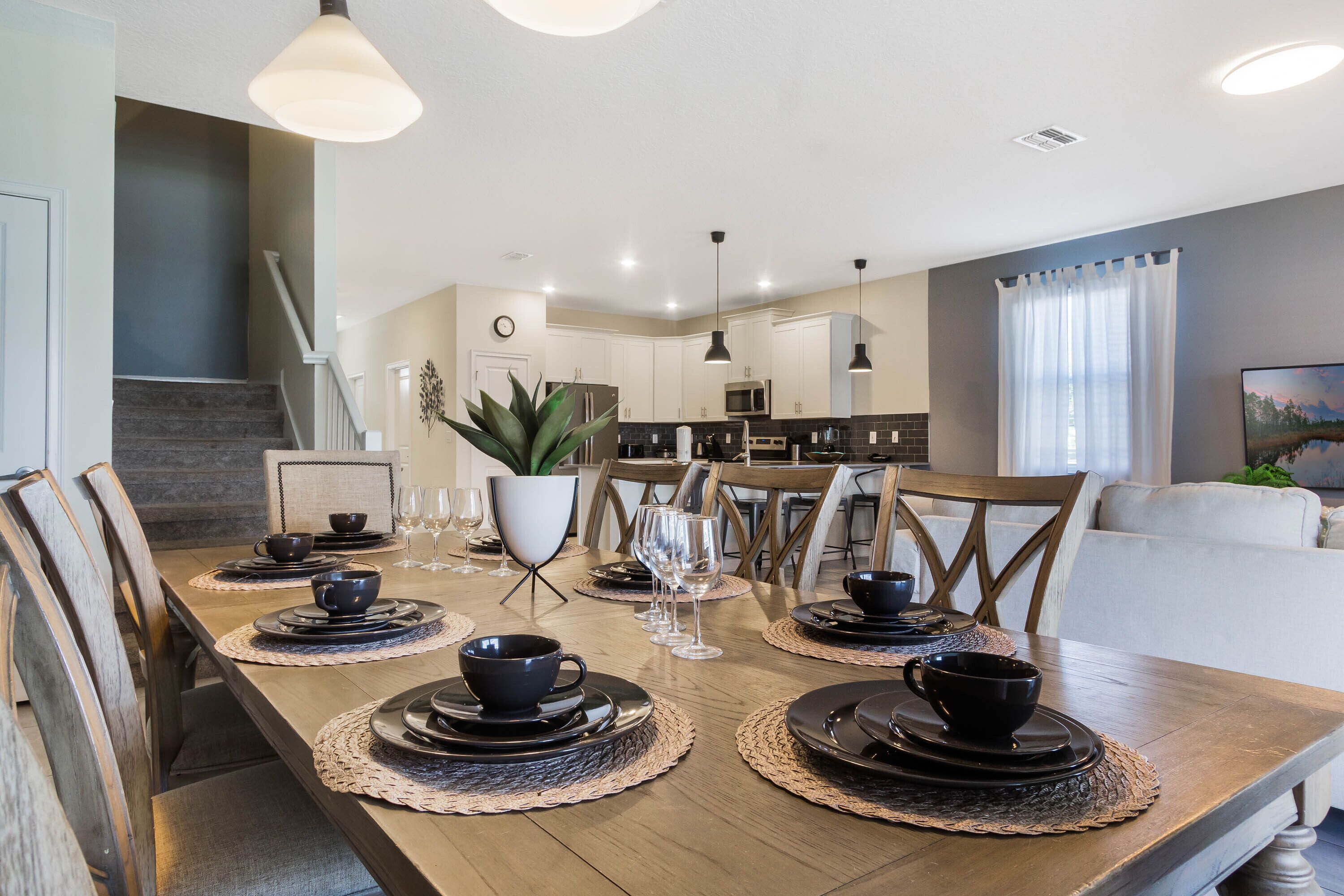 Une salle à manger avec une table en bois dressée pour quatre personnes, entourée de chaises. On aperçoit une cuisine au fond.