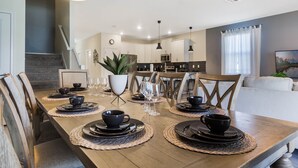 Un comedor con una mesa de madera dispuesta para cuatro personas, rodeada de sillas. Se observa una cocina al fondo.