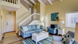 A spacious living room with a staircase, a blue sofa, a white coffee table, and a patterned rug. - 1607 Port Villas - Gorgeous views of Braddock Cove (Hilton Head Island)