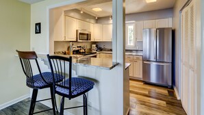 A kitchen with white cabinets, a stainless steel refrigerator, and a microwave. There are two bar stools with blue patterned seats. - 1607 Port Villas - Gorgeous views of Braddock Cove (Hilton Head Island)