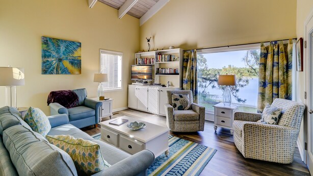 A spacious living room with a large window, a bookshelf, and a television. - 1607 Port Villas - Gorgeous views of Braddock Cove (Hilton Head Island)