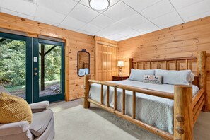 A wooden cabin bedroom with a bed, a nightstand, a lamp, and a view of the outdoors through a door.