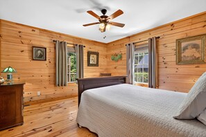 Una habitación con paredes de madera, un ventilador de techo, una cama con colcha blanca, una mesita de noche con lámpara y cuadros en las paredes.
