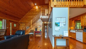 A spacious wooden cabin interior with a central staircase, a living area with a sofa, and a kitchen area. - Secluded Log Cabin on Wildlife Preserve | Mt. Mitchell Views, Game Loft & 3 Decks (Old Fort)