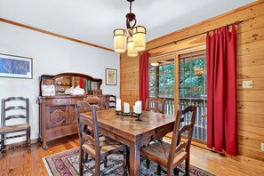 A dining room with a wooden table, chairs, a chandelier, and a view of the outdoors through a window with red curtains. - Secluded Log Cabin on Wildlife Preserve | Mt. Mitchell Views, Game Loft & 3 Decks (Old Fort)