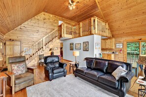 A spacious living area with a high ceiling, wooden walls, a leather sofa, a black armchair, a wooden staircase, and a kitchen area. - Secluded Log Cabin on Wildlife Preserve | Mt. Mitchell Views, Game Loft & 3 Decks (Old Fort)