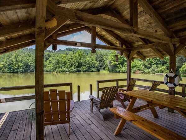 Una terraza de madera con mesas y sillas de picnic con vista a un lago tranquilo rodeado de bosque.
