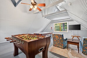 Una habitación con mesa de futbolín, ventilador de techo, televisor y ventana con vista a árboles.