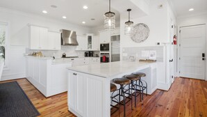 Cozinha moderna com armários brancos, uma ilha grande com banquetas, eletrodomésticos de aço inoxidável e iluminação pendente.