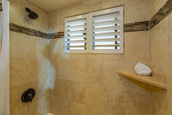 Un espace douche avec des carreaux de couleur beige, une fenêtre avec des stores, une serviette posée sur une étagère et un pommeau de douche.