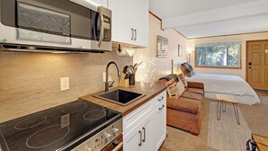Una cocina moderna con estufa integrada, microondas y lavabo. Hay una encimera de madera y armarios blancos. En el área de estar contigua se ve un sofá de cuero marrón y una pequeña mesa de madera.