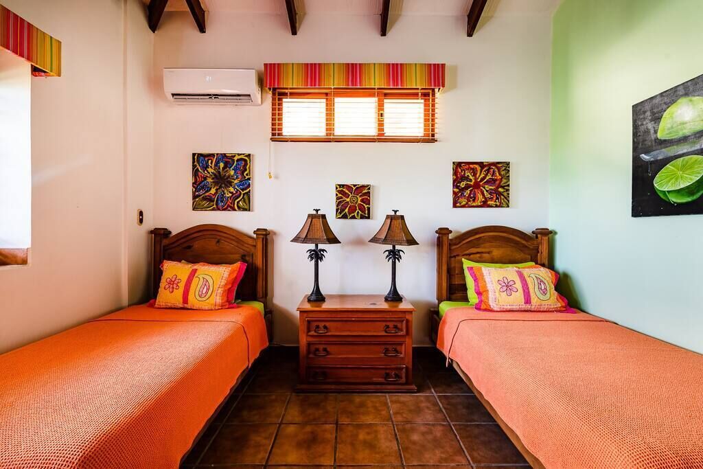 A room with two beds, each with a wooden headboard and orange bedding. There are two nightstands with lamps, a small wooden dresser, and a window with blinds.