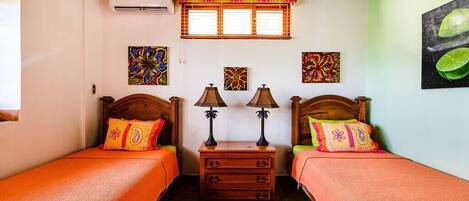 A room with two beds, each with a wooden headboard and orange bedding. There are two nightstands with lamps, a small wooden dresser, and a window with blinds.