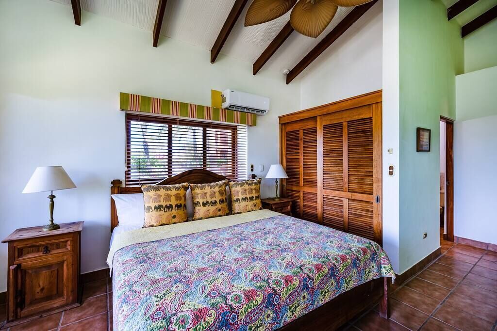 A bedroom with a bed, wooden headboard, two bedside tables with lamps, a window with blinds, and a wooden wardrobe.