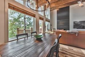 A dining area with a wooden table, chairs, and a green bowl. There is a large window with a view of trees and a balcony.
