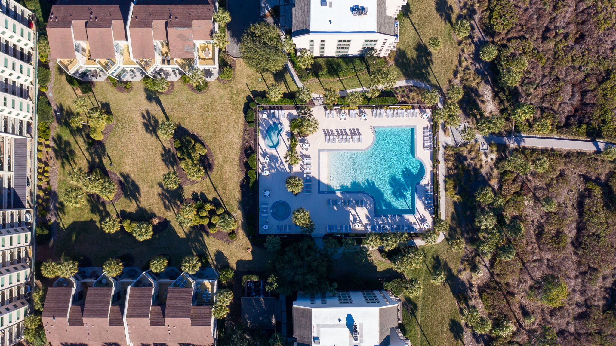 An aerial view of a resort with a central swimming pool, multiple buildings, and landscaped areas with palm trees.