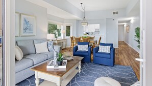 A spacious living room with a grey sofa, blue armchairs, a coffee table, and a dining area with wicker furniture.