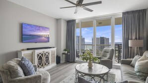 A spacious living room with a large window offering a city view, a ceiling fan, and a flat-screen TV mounted on the wall. - St. Lucia 904 ~  9th Floor ~ Amazing Views! (Destin)