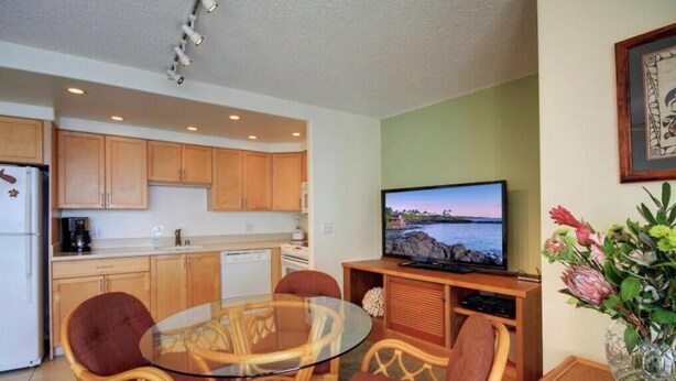 Un espacio habitable compacto con kitchenette, una mesa de comedor de vidrio, sillas de mimbre, un televisor de pantalla plana y un florero con flores.