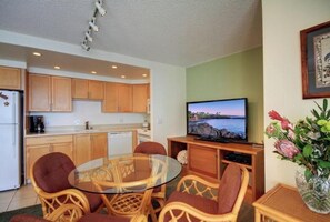 A compact living space with a kitchenette, a glass-top dining table, wicker chairs, a flat-screen TV, and a vase with flowers.
