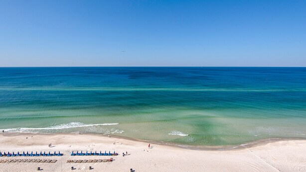 Une plage à l’eau turquoise et limpide, bordée d’un rivage sablonneux et d’une rangée de parasols de plage bleus.
