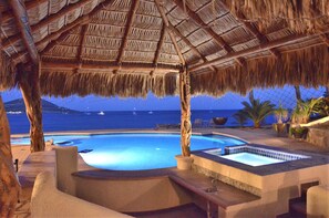 A pool area with a thatched roof, a hot tub, and a view of the sea.