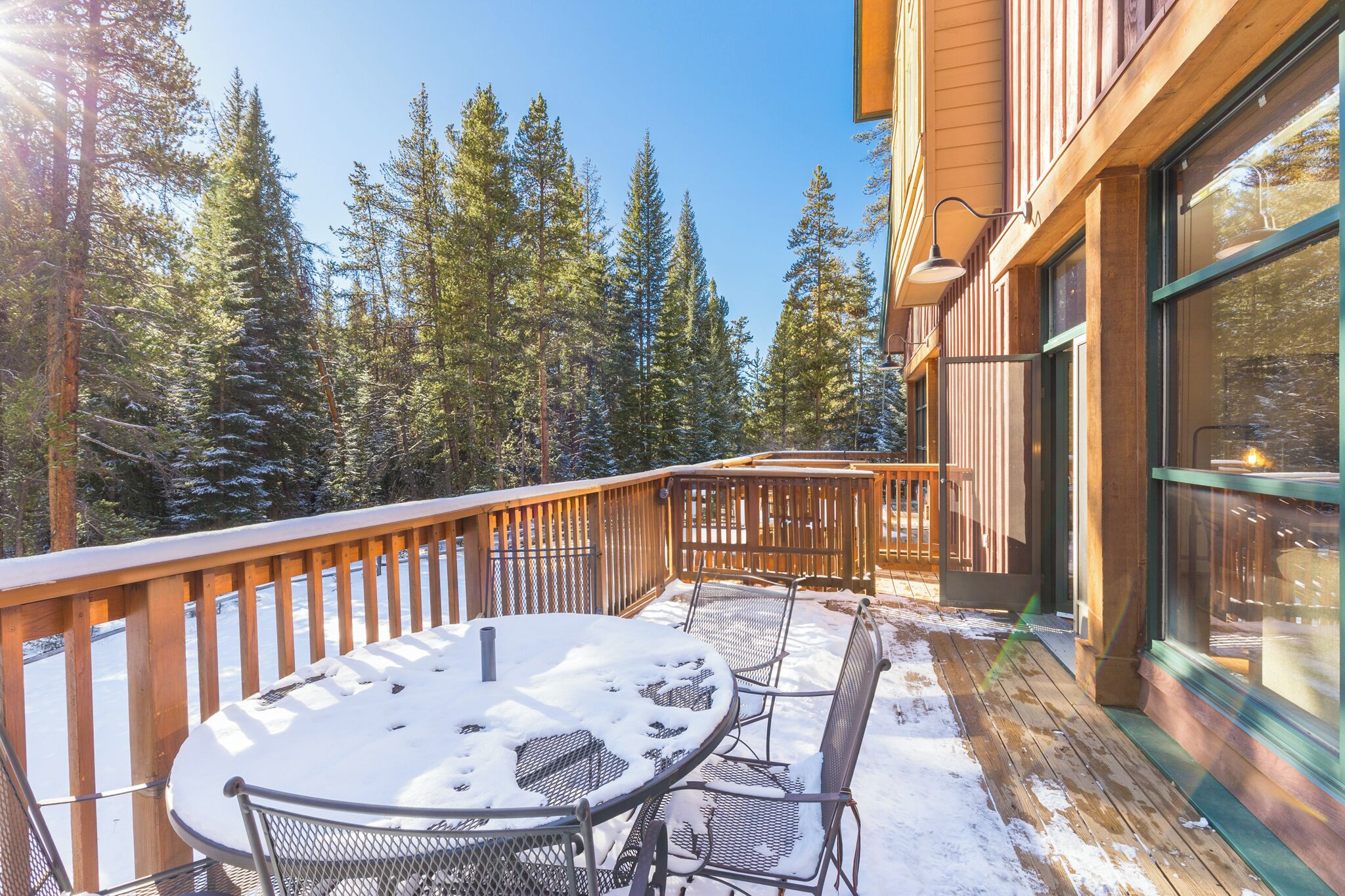 A wooden deck with a snow-covered table and chairs, surrounded by tall pine trees.