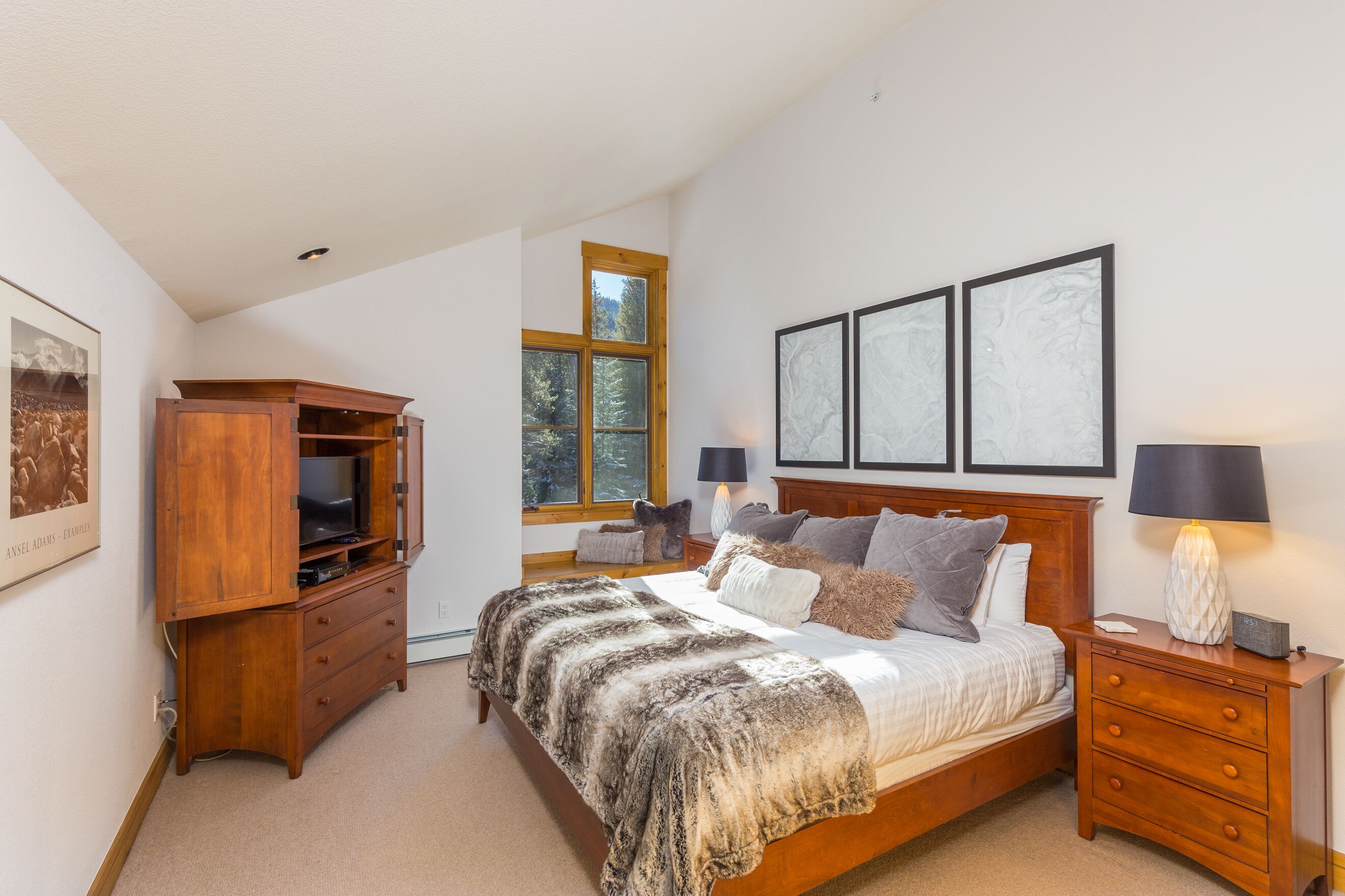 A bedroom with a bed, wooden nightstands, a TV cabinet, and a window with a view of trees.