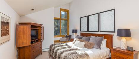 A bedroom with a bed, wooden nightstands, a TV cabinet, and a window with a view of trees.