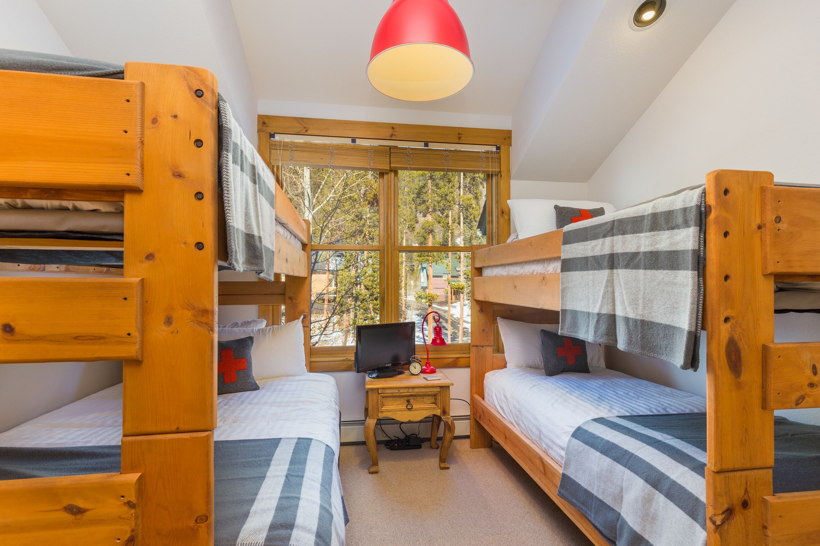 A dormitory room with bunk beds, a small desk with a computer, and a window with a view of trees.