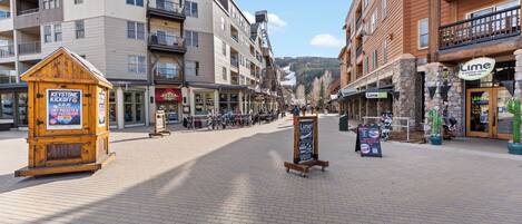 Une zone piétonne dallée, bordée d’immeubles à plusieurs étages, avec un kiosque en bois et un panneau d’affichage.