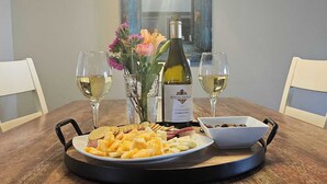 Een eettafel met een bord kaas, crackers en olijven, twee glazen witte wijn, een fles Chardonnay en een vaas met bloemen.