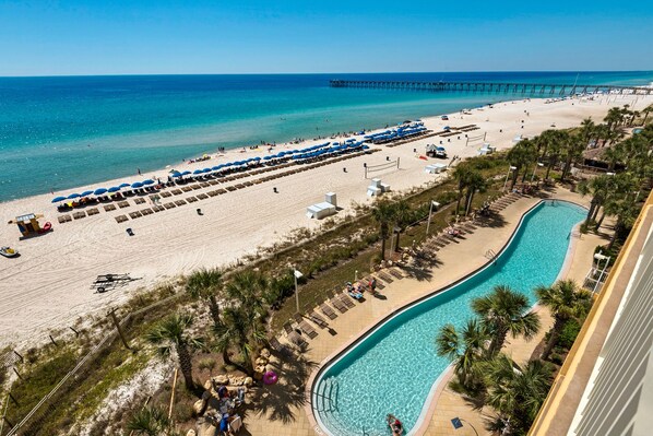 A beach resort with a swimming pool, beach umbrellas, and a pier in the background.