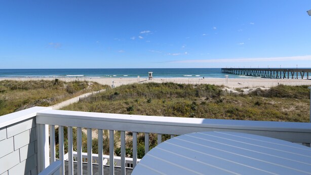 A beach view from a balcony with a wooden railing, a sandy beach, and a pier extending into the water.