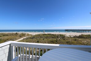 A beach view from a balcony with a wooden railing, a sandy beach, and a pier extending into the water.