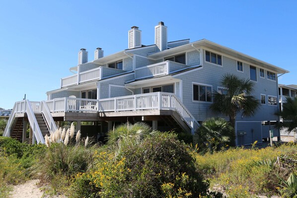 A multi-story beachfront house with a balcony, a staircase leading to the upper floor, and a sandy path in the foreground.