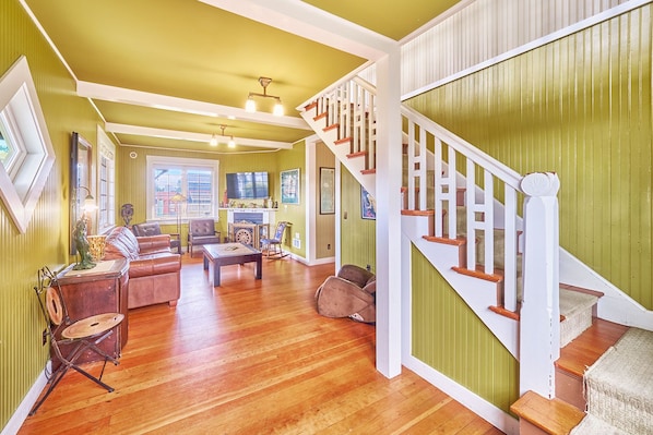 A spacious living room with a wooden floor, a staircase with white railings, and a green wall.