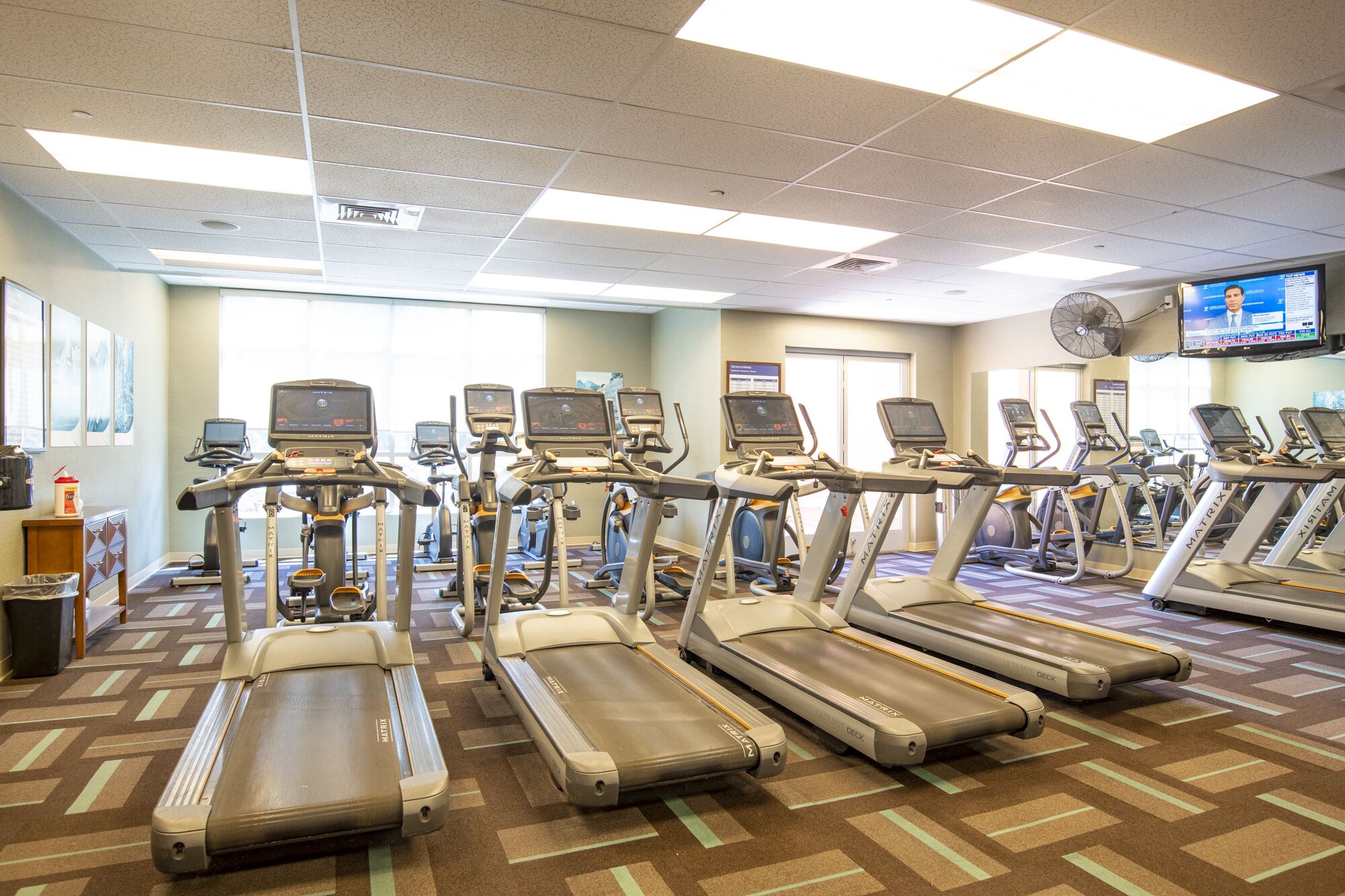 A gym with multiple treadmills and elliptical machines, a television mounted on the wall, and a patterned carpet.