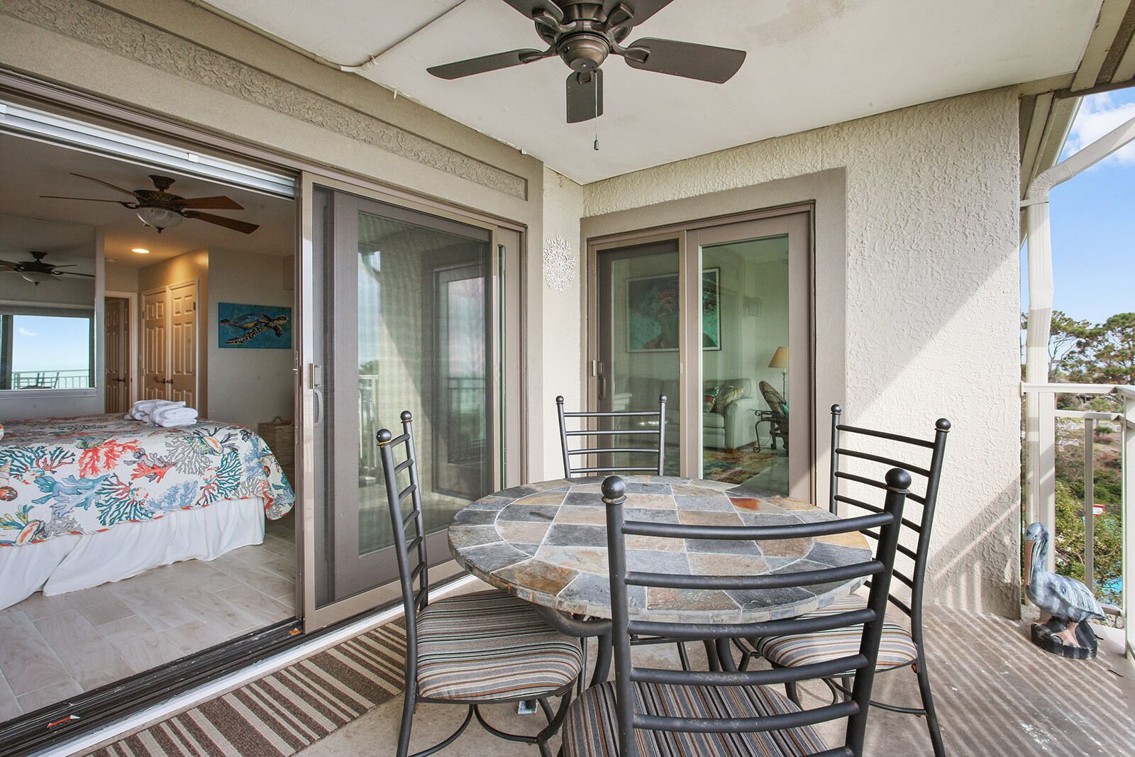 A balcony with a table and chairs, a ceiling fan, and a sliding glass door leading to a bedroom with a bed and a painting.