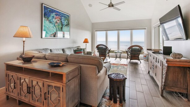 A spacious living area with a large sofa, a wooden cabinet, and a television. There is a ceiling fan, a window with a view of the ocean, and a table with chairs. - Shorewood 504- Ocean Front Top Floor Spectacular Views! (Hilton Head Island)