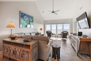A spacious living area with a large sofa, a wooden cabinet, and a television. There is a ceiling fan, a window with a view of the ocean, and a table with chairs.